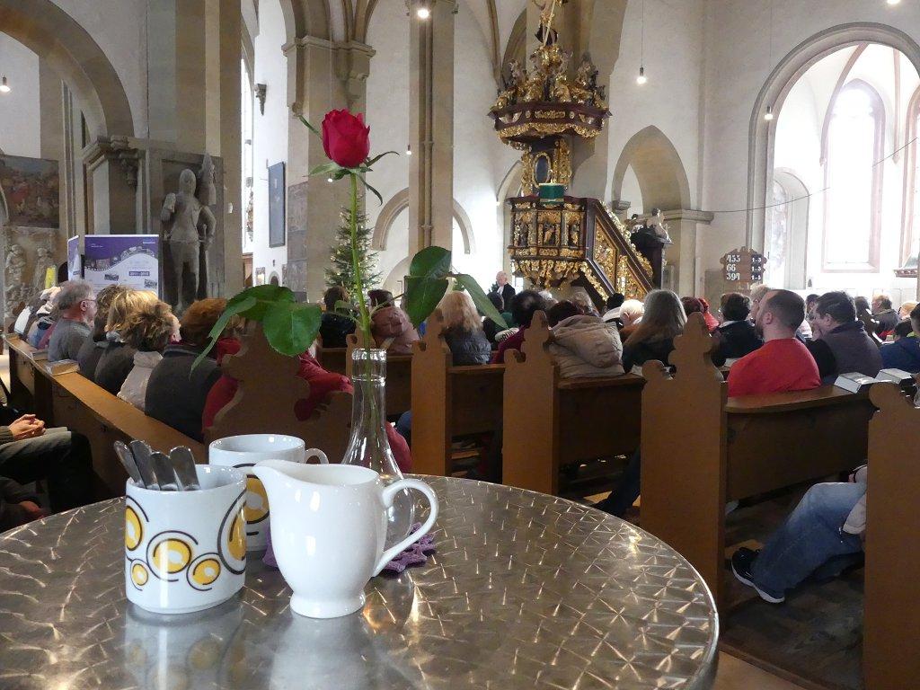 Eine Tasse mit Vesperkirche-Logo und ein Milchkännchen sowie eine Rose stehen auf einem Stehtisch im Vordergrund. Dahinter ist die Gottesdienstgemeinde zu erkennen.