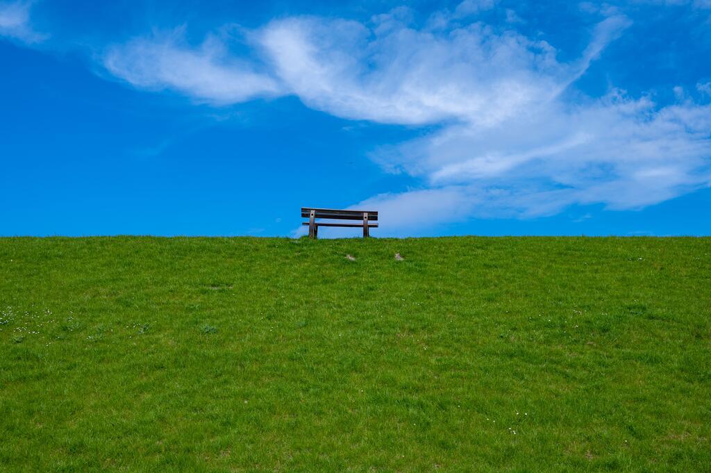 Auf einem grünen Deich steht eine alte Bank. Darüber sieht man nur blauen Himmel. 