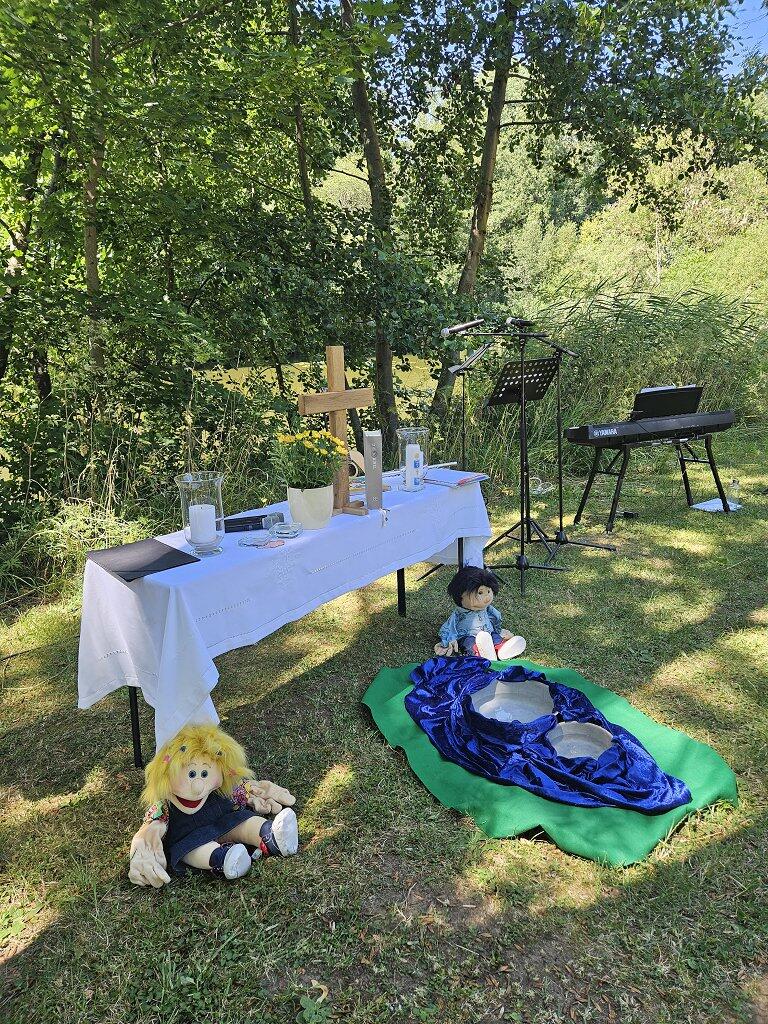 Ein Altar im Freien am See, davor sitzen zwei Handpuppen