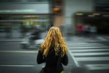 Eine junge Frau mit langen Haaren von hinten. Sie blickt auf eine unscharf dargestellte Straße, auf der anderen Seite Läden, rechts ein Zebrastreifen, alles ganz verschwommen.