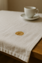 Das Bild zeigt eine weiße Stofftischdecke auf einem hellen Holztisch. Im Zentrum befindet sich ein kleiner, dunkler Kaffeefleck, der sich leicht in die Fasern ausbreitet. Im Hintergrund, unscharf und nur angedeutet, steht eine leere Kaffeetasse auf einem Unterteller. Natürliches, weiches Licht fällt schräg von der Seite ein und betont die ruhige, fast alltägliche Szene. Die Komposition wirkt still, fast meditativ – mit einem Hauch von Ironie durch den kleinen „Makel“ im Mittelpunkt.