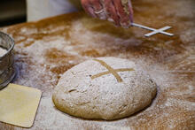 In einer Bäckerei wird Brot gebacken für die Aktion "5.000 Brote - Brot für die Welt". Im Vordergrund ein Brot und eine Hand, die ein Kreuz hält, um das Kreuz als Motiv auf das Brot zu bringen.