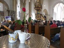 In der Vesperkirche Schweinfurt stehen Milchkännchen und Tische bereit. Die Menschen sitzen noch zum Gottesdienst in den Bänken.